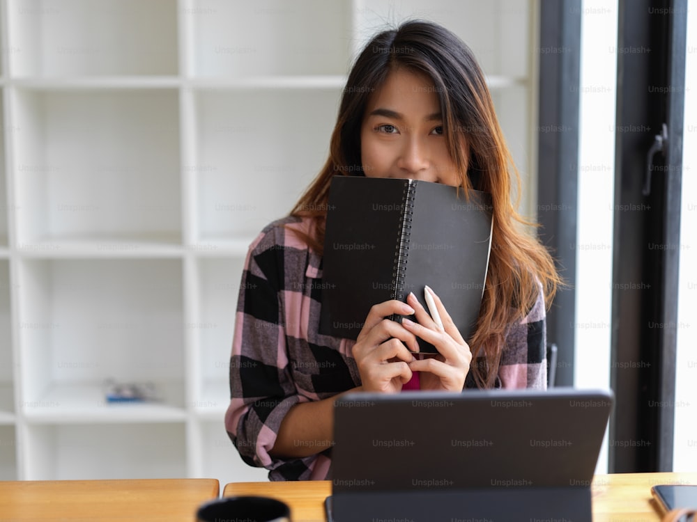 Young beautiful woman cover her mouth with notebook in modern workspace