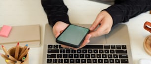 Top view of female hands using smartphone while working with laptop and stationery in home office room