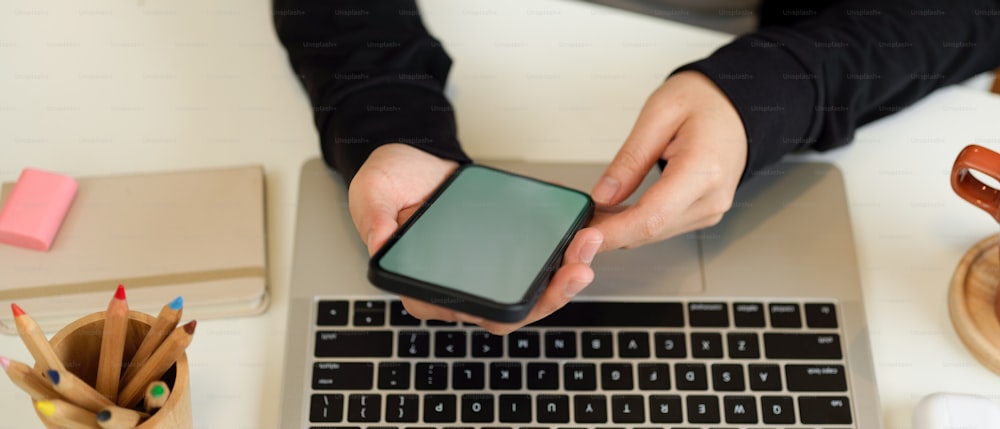 Draufsicht auf weibliche Hände mit Smartphone bei der Arbeit mit Laptop und Schreibwaren im Home-Office-Raum