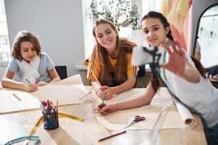 Adorable female child recording video with smartphone and smiling while little seamstresses drafting sewing patterns in tailoring atelier