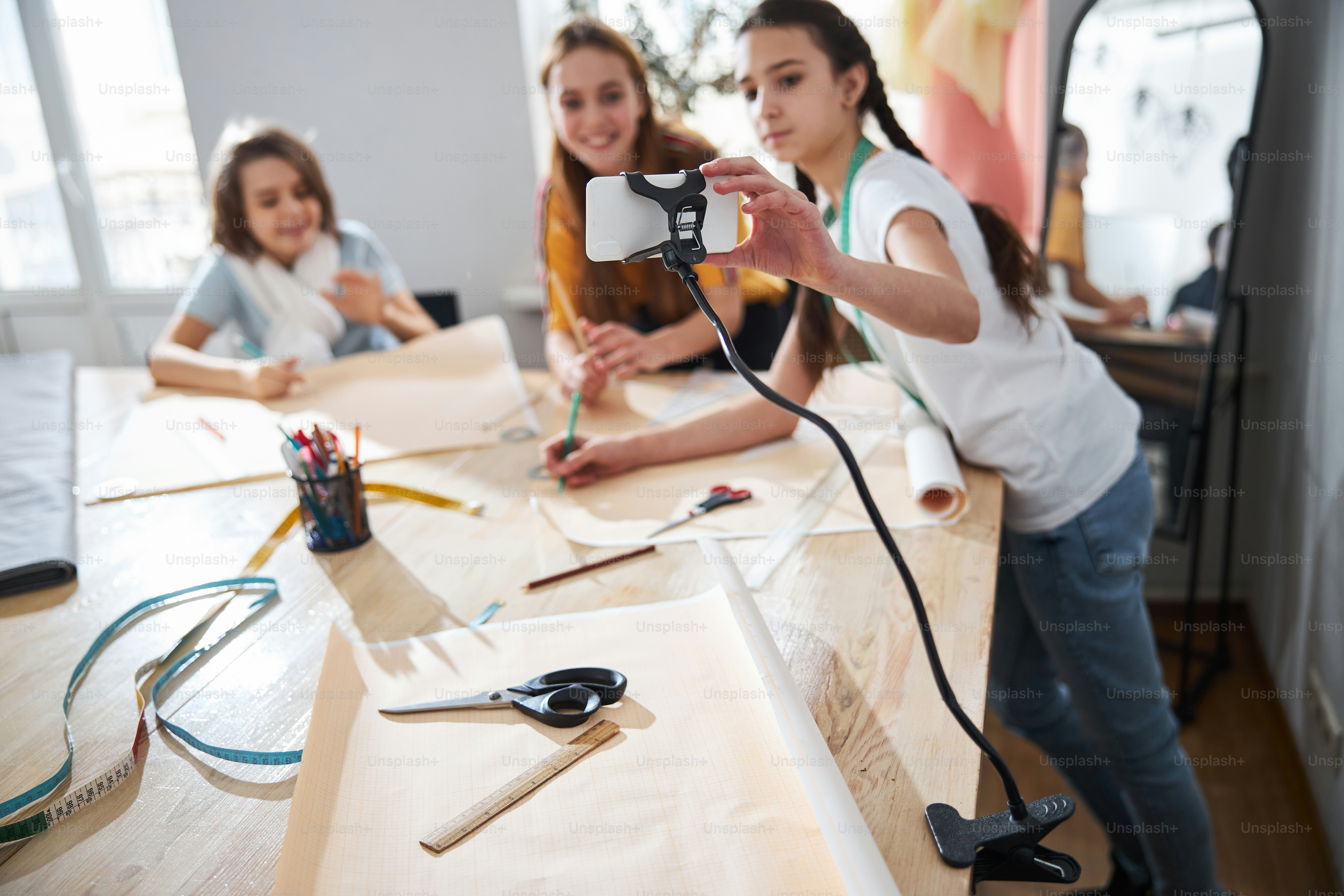 Cute female child recording video with smartphone while little seamstresses drafting sewing patterns in tailoring atelier