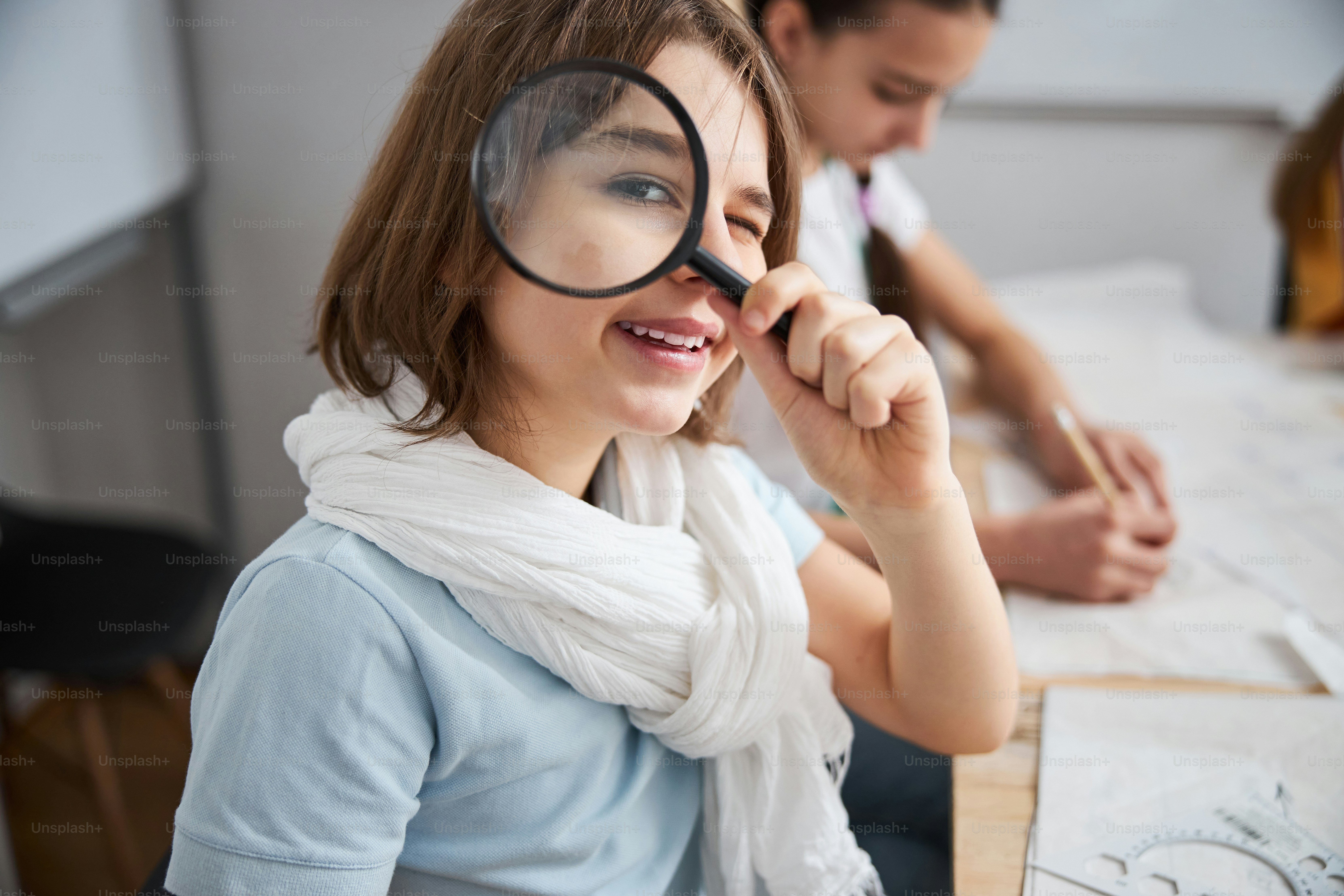 Adorable female child holding loupe and smiling while sitting at the table and drawing sewing patterns