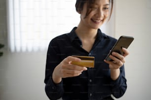 Mulher jovem sorridente segurando cartão de crédito pagando contas on-line ou fazendo compras on-line com telefone inteligente.