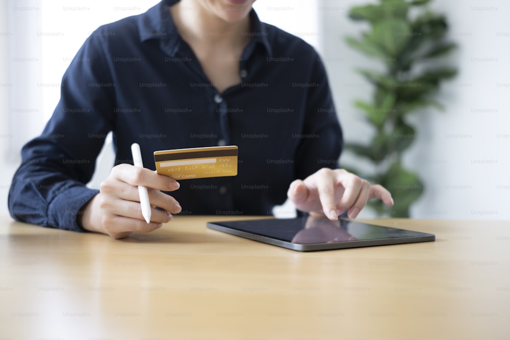 신용 카드를 들고 디지털 태블릿을 사용하여 온라인으로 청구서를 지불하거나 온라인 쇼핑을 하는 젊은 여성의 잘린 사진.