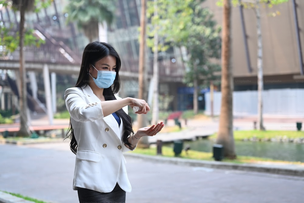 Junge Frau in Schutzmaske trägt Alkoholspray auf, um ihre Hände im Freien zu reinigen und zu desinfizieren.