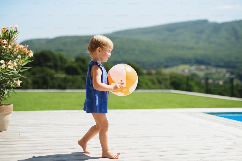 Side view portrait of small toddler girl with ball walking outdoors in backyard garden.