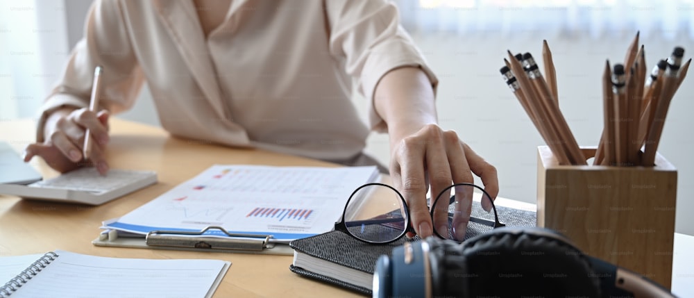 Foto recortada de una mujer de negocios que recoge sus gafas y usa la calculadora para calcular en el escritorio de la oficina.