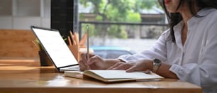 Cropped shot of young woman using computer laptop and writing information on notebook.