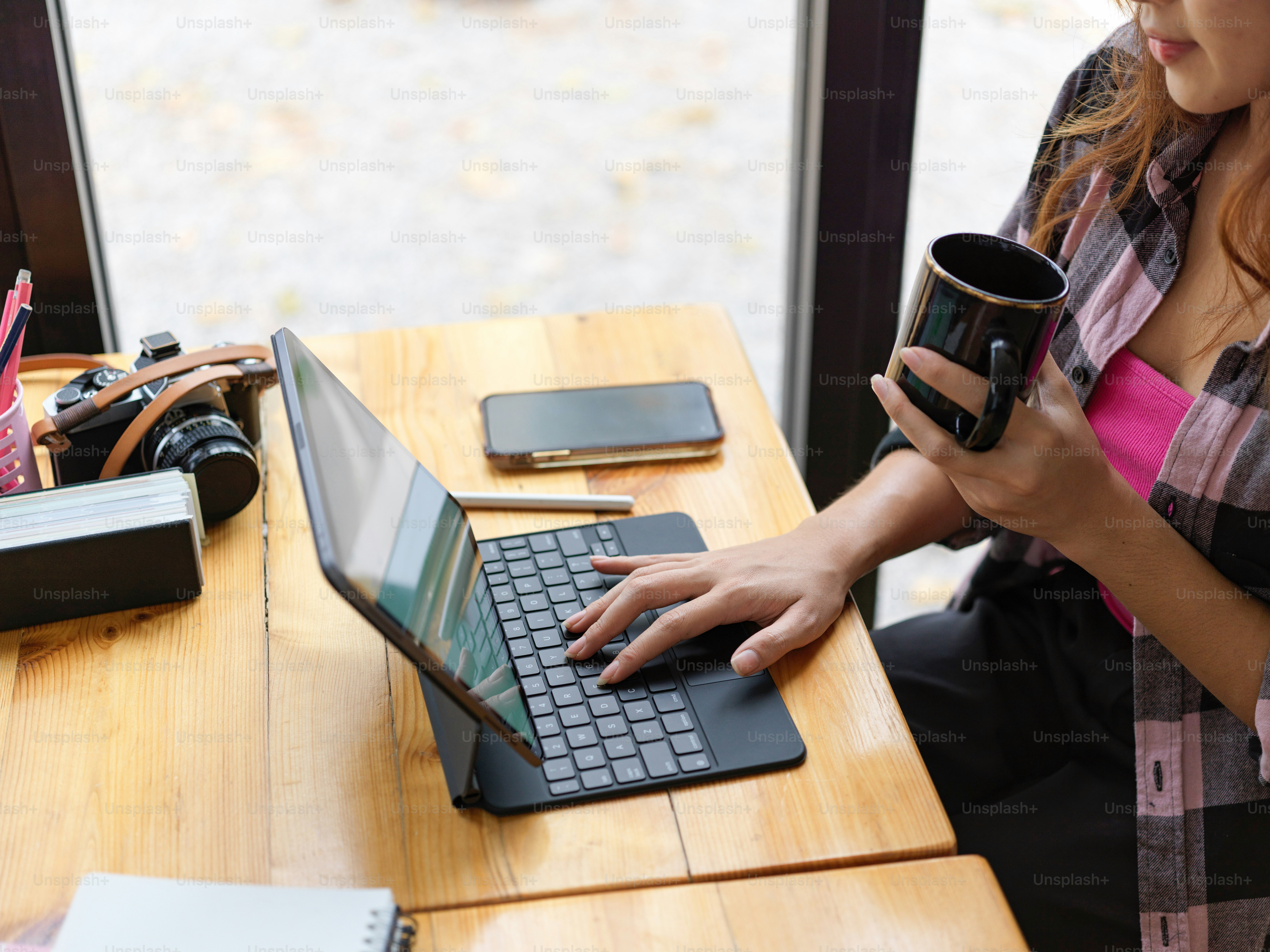 Tampilan samping pekerja lepas wanita muda memegang cangkir kopi dan bekerja dengan tablet digital di atas meja kayu di kafe
