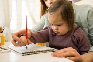 Nahaufnahme des weiblichen Kindes mit Down-Syndrom, das mit Farben auf Papier zeichnet, während es am Tisch an ihrem Arbeitsplatz sitzt. Mutter hilft beim Zeichnen zu ihrer Tochter. Archivfoto