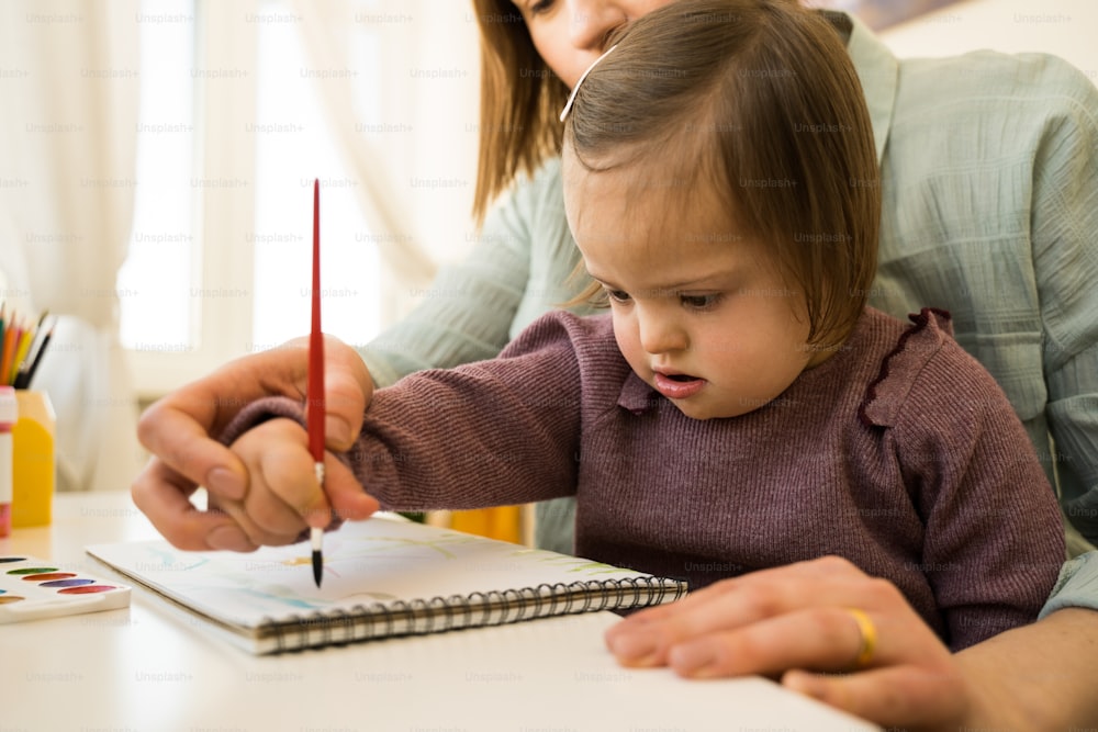 Close up view of the female kid with down syndrome drawing with paints on paper while sitting at the table at her workplace. Mother helping drawing to her daughter. Stock photo