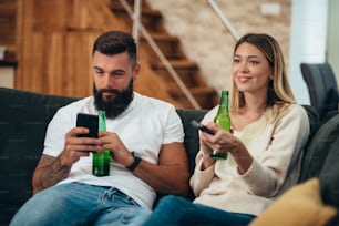 Jovem casal relaxando em casa e bebendo cerveja enquanto usa um smartphone