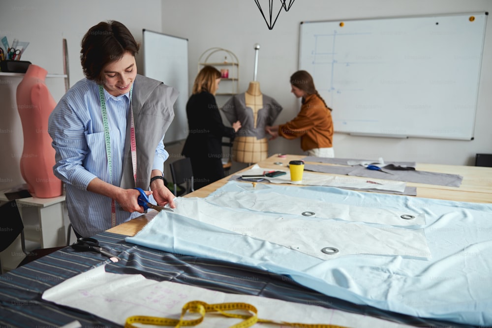 Glad fashion designer operating with dressmaking shears and cutting fabric on a large table along the pattern edge
