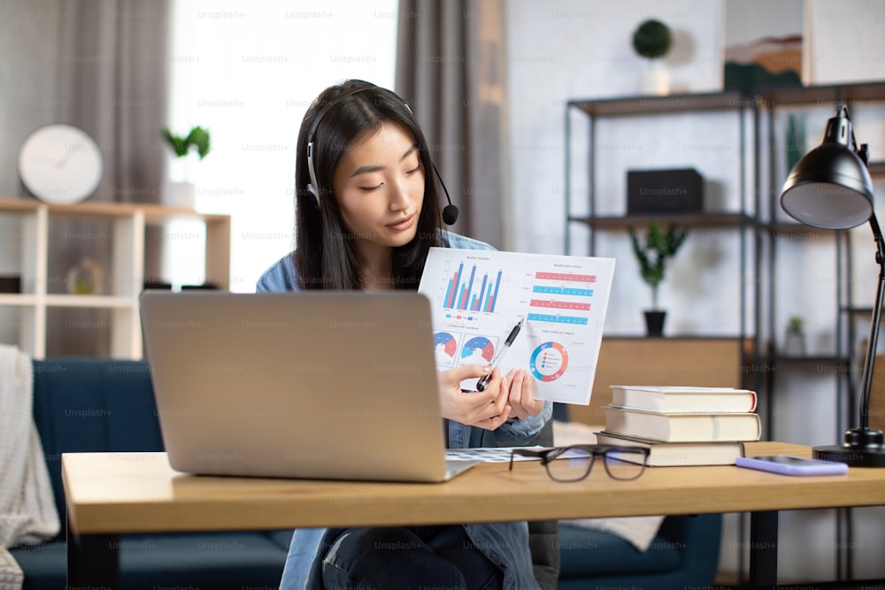 Serious asian woman in headset having video conference on laptop while working from home. Attractive Korean girl student sitting at desk and showing graphs and charts to her colleagues via laptop