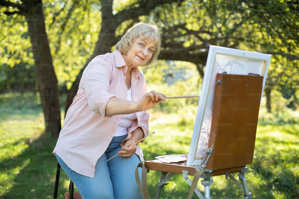Mulher aposentada caucasiana feliz, vestindo camisa leve e jeans, pintando em tela e se divertindo no belo jardim verde ou parque em dia ensolarado. Arte, hobby e conceito de aposentadoria.
