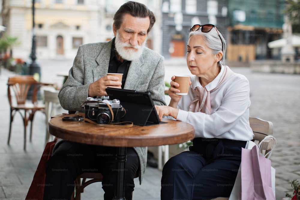 Mature man and woman in stylish outfits using digital tablet while drinking coffee on cafe terrace. Happy senior couple relaxing outdoors with modern technology.
