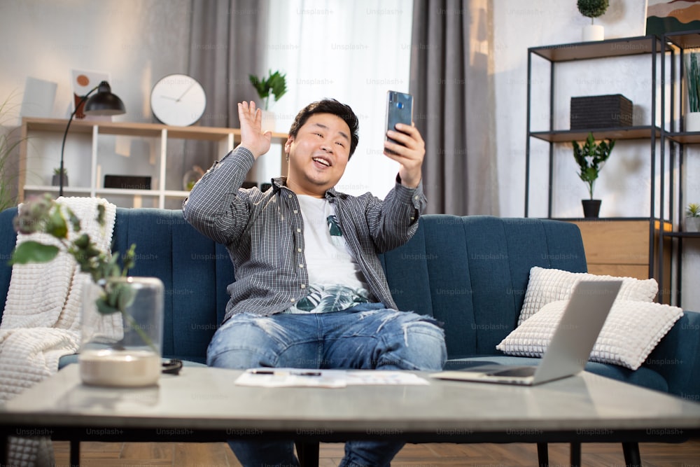 Smiling asian man in casual clothes sitting on couch and using modern smartphone for video conversation. Corporate communication and remote work.
