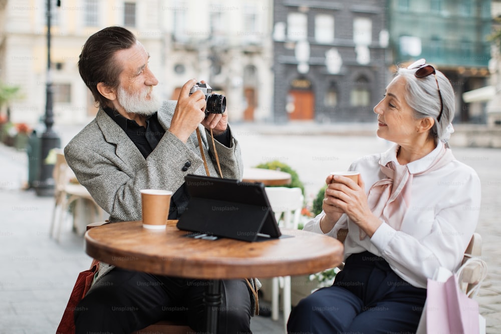Mature bearded man using retro camera for taking photo of his pretty lovely wife. Happy couple in stylish wear relaxing on cafe terrace with tablet and drinking coffee.