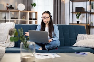 Giovane donna fiduciosa in cuffia seduta sul divano e in videoconferenza sul laptop. Libera professionista che conduce riunioni di lavoro online rimanendo a casa.
