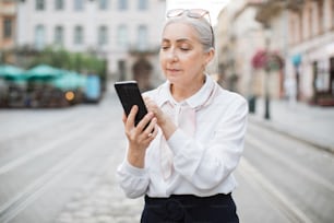 Mujer mayor bonita que usa un teléfono inteligente moderno para videollamadas. Dama de cabello gris con un atuendo elegante que camina por la calle de la ciudad y habla en línea.