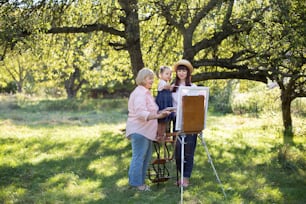 Bellissimo scatto primaverile estivo di famiglia felice di tre generazioni, bambina piccola, giovane madre e nonna matura, che trascorre del tempo all'aperto e dipinge l'immagine sul cavalletto. Tempo libero estivo in famiglia.