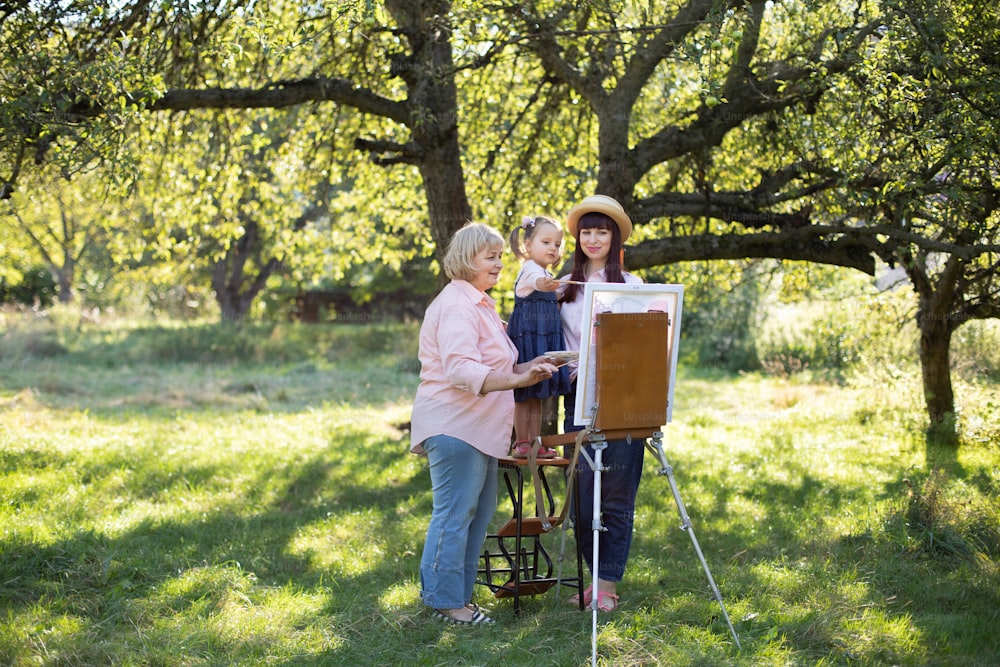Beautiful summer spring shot of happy three generations family, little kid girl, young mother and mature grandmother, spending time outdoors and painting picture on easel. Summer family leisure.
