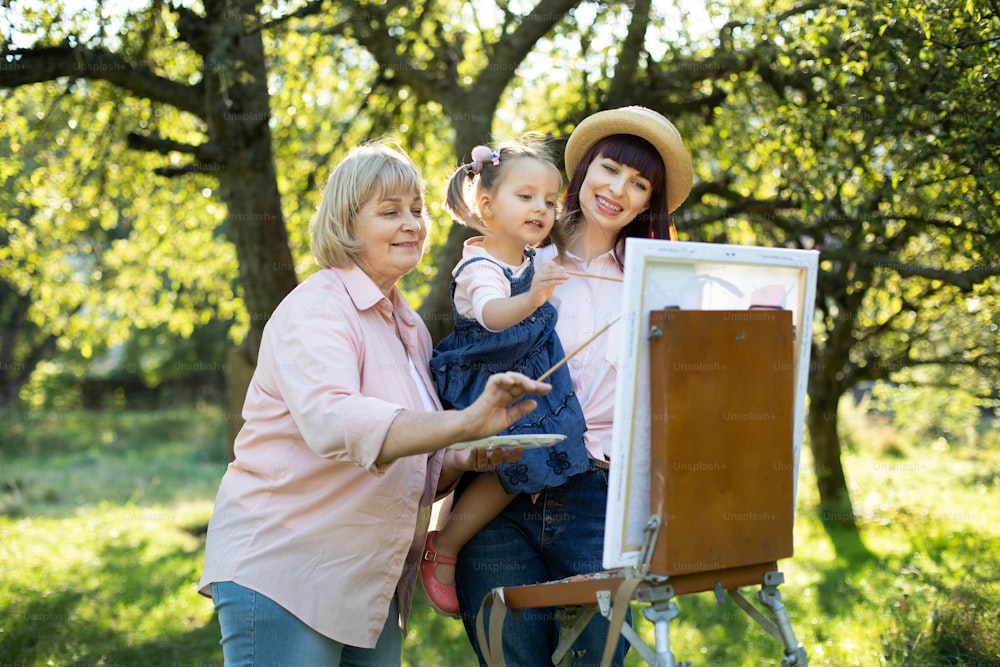 Bella famiglia caucasica di tre generazioni, nonna, madre e bambina carina che si trova fuori nel parco primaverile estivo e dipinge un'immagine su cavalletto di legno.