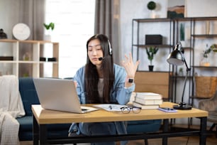 헤드셋을 쓴 예쁜 윤 아시아 여성은 노트북으로 화상 채팅을 하는 동안 말하고 몸짓을 하고, 집에서 밝고 현대적인 방에서 온라인으로 일하거나 공부합니다. 판데미 기간 동안의 원격 작업 및 교육