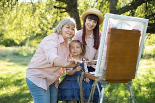 Famiglia felice, tre generazioni di donne. Pittura di famiglia all'aperto. Bambina carina che si diverte con la mamma e la nonna, dipingendo insieme un quadro su un cavalletto al giardino primaverile.