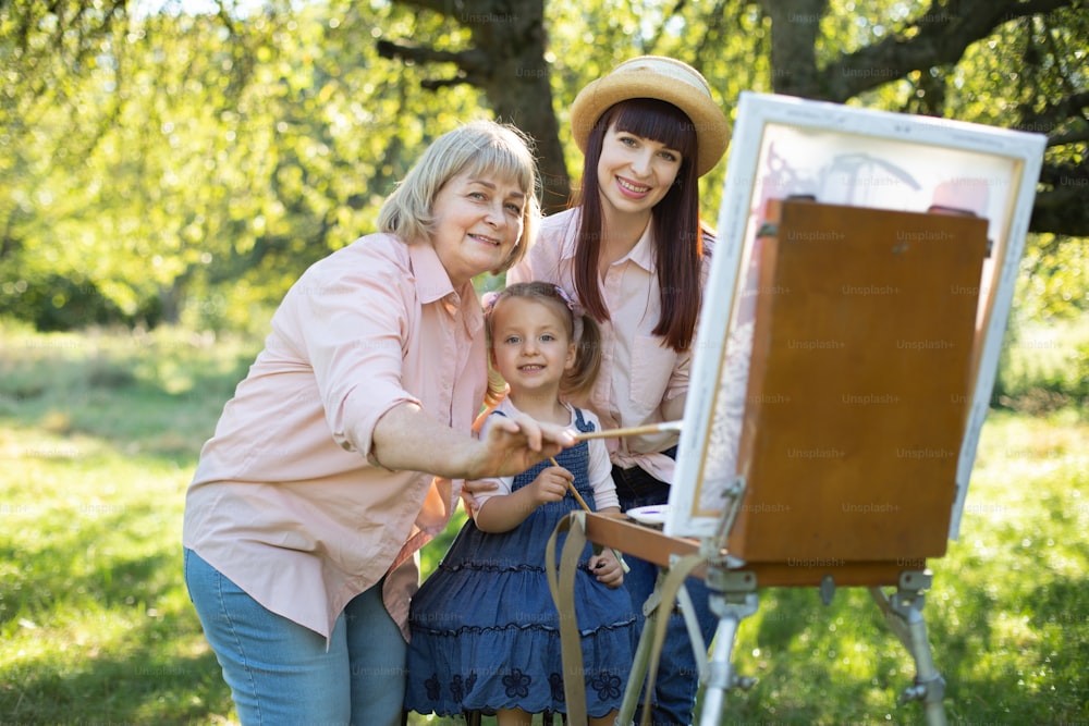 Famille heureuse trois générations de femmes. Peinture de famille à l’extérieur. Jolie petite fille s’amusant avec sa mère et sa grand-mère, peignant ensemble une image sur un chevalet au jardin de printemps.