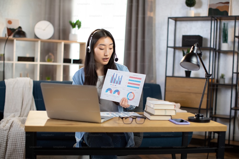 Professional young Korean woman presenting financial charts and graphs to colleagues through video call on laptop. Female freelancer or manager sitting at workplace and talking in headset.