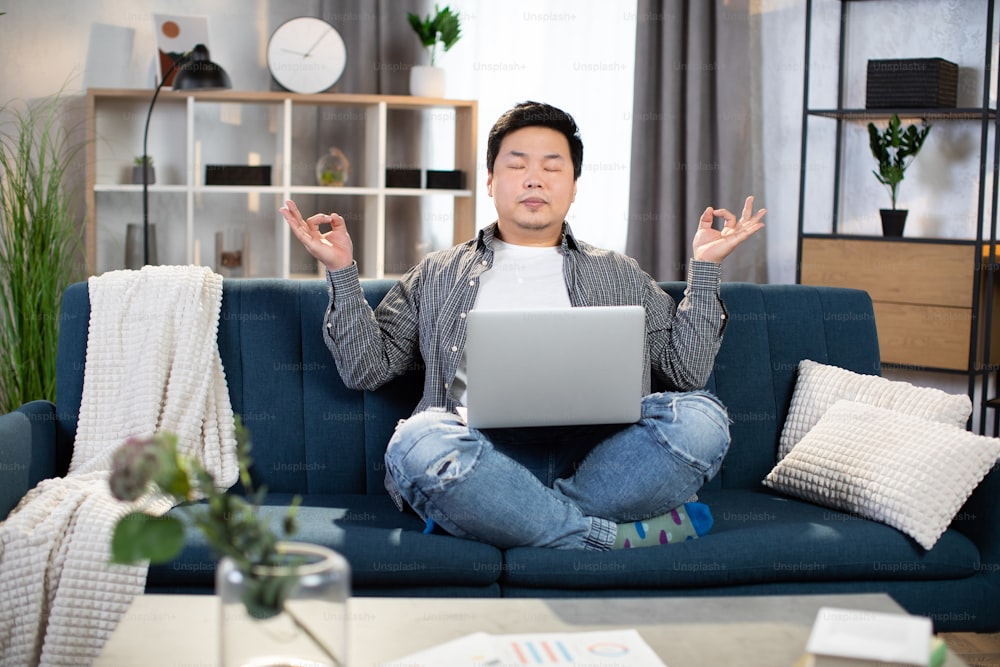 Hombre asiático relajado sentado en el sofá con una computadora portátil de rodillas y meditando con los ojos cerrados. Freelancer cansado tomando un descanso durante el proceso de trabajo en casa.
