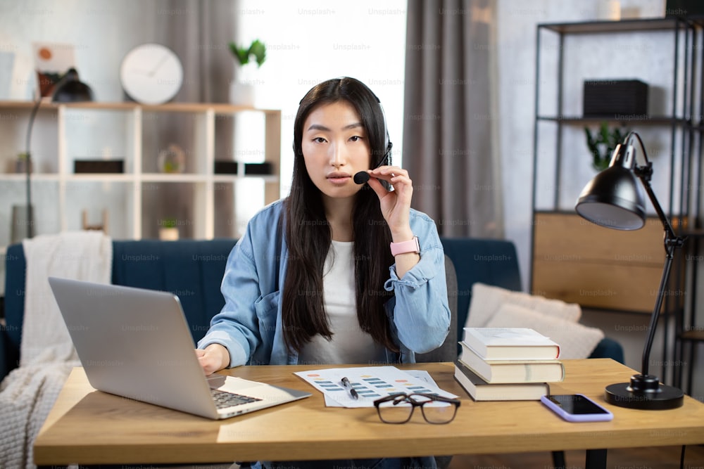 Lavoro e apprendimento a distanza. Giovane studentessa asiatica che utilizza l'auricolare e il laptop per la conversazione video, studiando a casa. Libera professionista seduta al tavolo e che prende appunti durante la chat video.