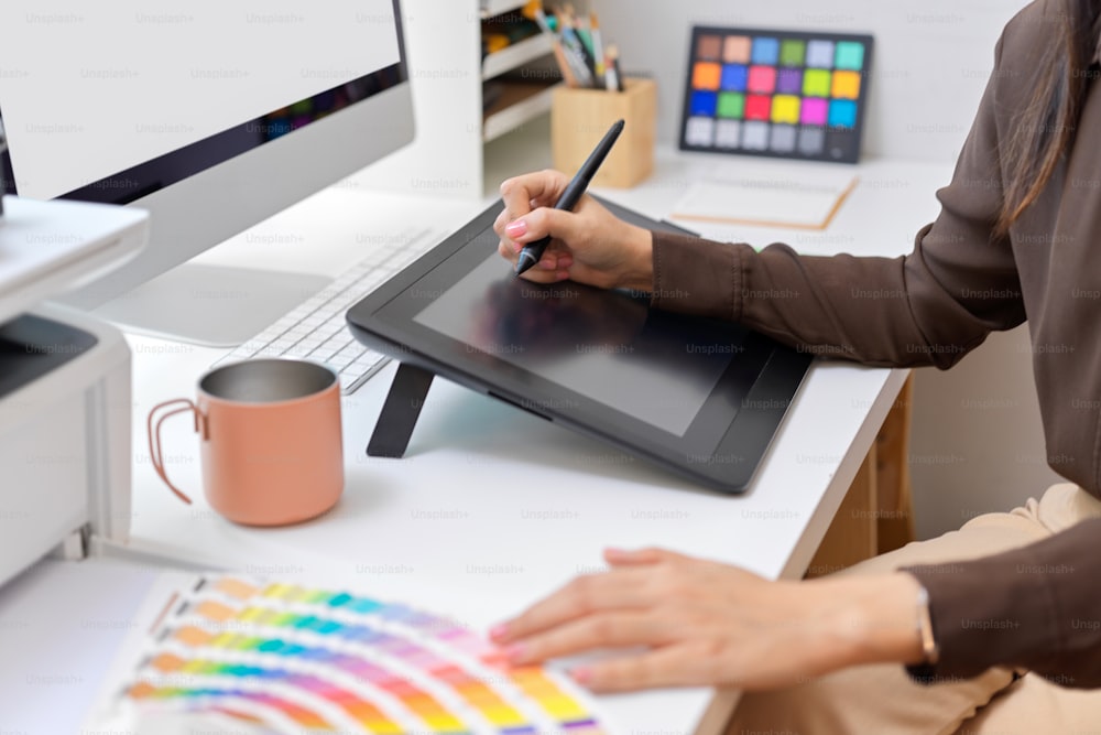 Vista lateral da mão do designer feminino que trabalha com o tablet de desenho e a amostra de cor na mesa do computador