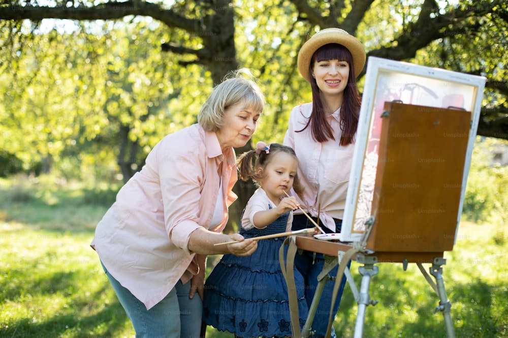 Familie von drei weiblichen Generationen im Freien. Glücklicher Familienurlaub in der Sommernatur. Angenehme Großmutter, hübsche Mutter und süßes kleines Mädchen, die zusammen auf einer Staffelei im grünen Park zeichnen.