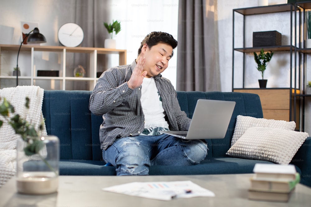 Asian man in casual outfit smiling and waving during video chat on modern laptop. Male freelancer sitting on couch and having online conference, working remotely from home