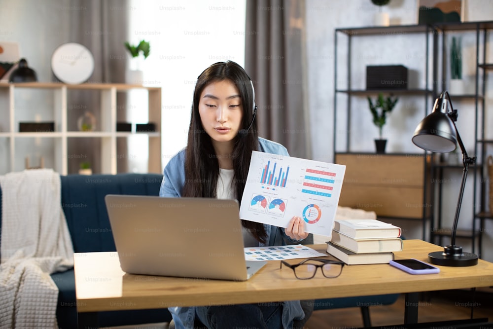 Mulher asiática apresentando relatório financeiro aos colegas através de chamada de vídeo em laptop. Freelancer do sexo feminino sentado no local de trabalho e falando em fone de ouvido.
