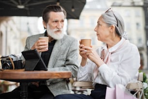 Stylish aged couple relaxing together on cafe terrace and enjoying hot coffee. Lovely wife and husband using digital tablet and retro camera outdoors.