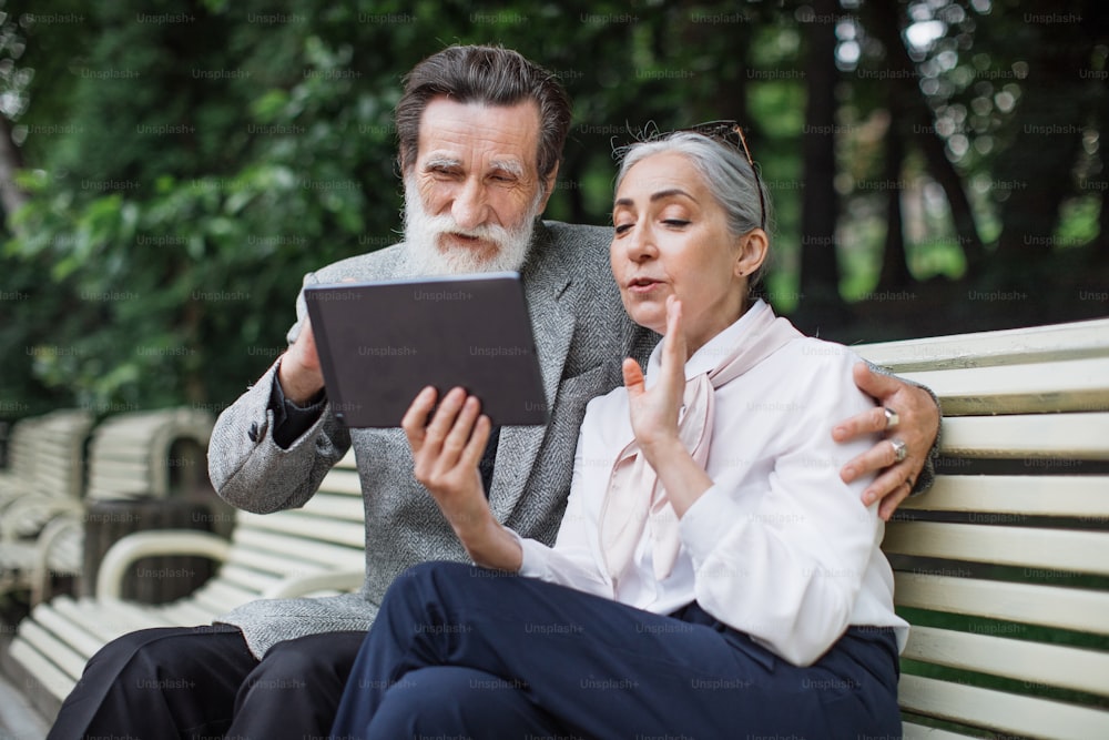 Beautiful caucasian family sitting on bench, looking on each other and holding digital tablet. Elder couple in stylish clothes relaxing together on fresh air.