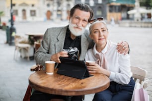 Angenehmes älteres Paar, das sich umarmt und lächelt, während es am Cafétisch mit digitalem Tablet und Retro-Kamera sitzt. Kaffeepause an der frischen Luft. Glücklicher Ruhestand.