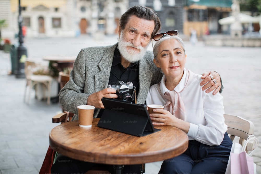 Pleasant senior couple hugging and smiling while sitting at cafe table with digital tablet and retro camera. Coffee break on fresh air. Happy retirement.