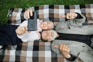 Vue de dessus d’un couple de personnes âgées souriant allongé sur une couverture douce et prenant un selfie sur un smartphone. Famille joyeuse se relaxant dans un parc vert et utilisant un gadget moderne.
