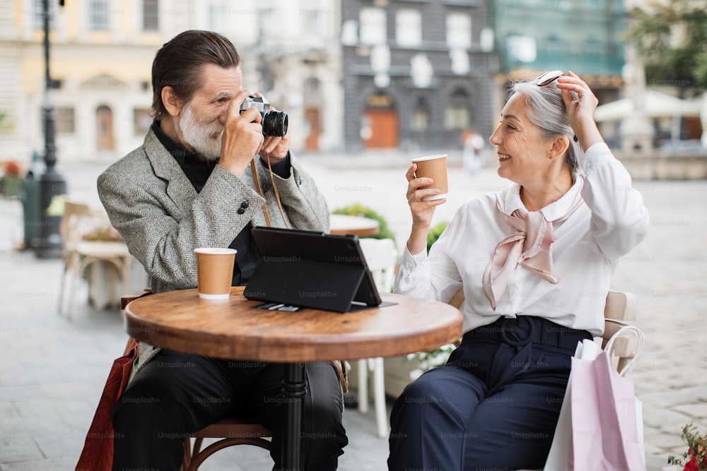 Mature bearded man using retro camera for taking photo of his pretty lovely wife. Happy couple in stylish wear relaxing on cafe terrace with tablet and drinking coffee.