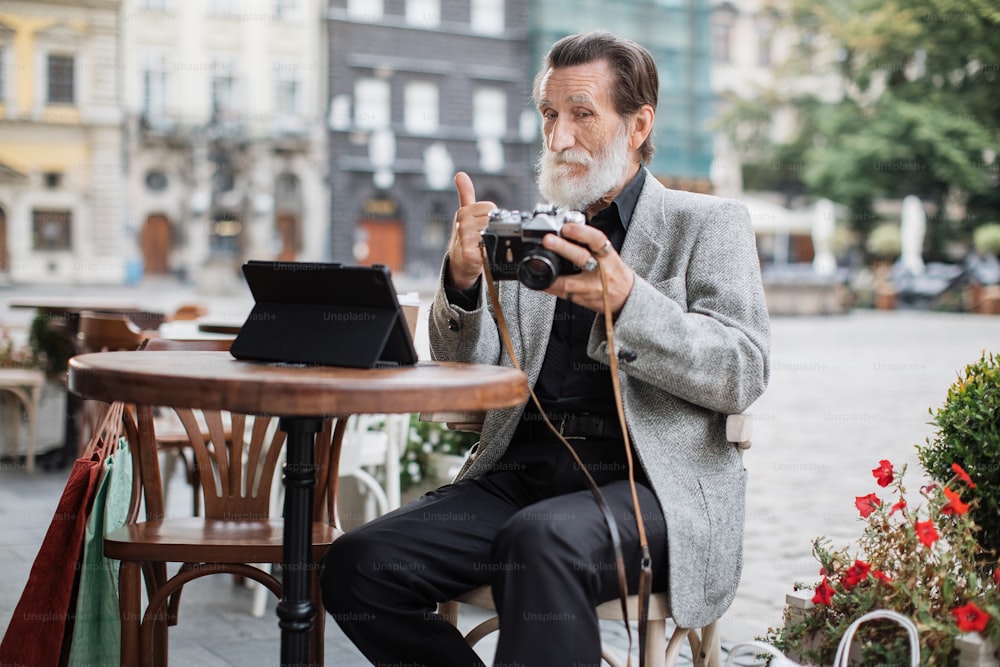 Hombre mayor seguro de sí mismo sentado en la terraza de la cafetería y usando una tableta digital con cámara retro. Jubilado barbudo revisando fotos y mostrando el pulgar hacia arriba.