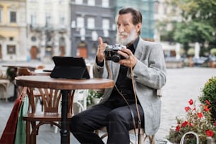 Confident senior man sitting at cafe terrace and using digital tablet with retro camera. Bearded pensioner reviewing pictures and showing thumb up.