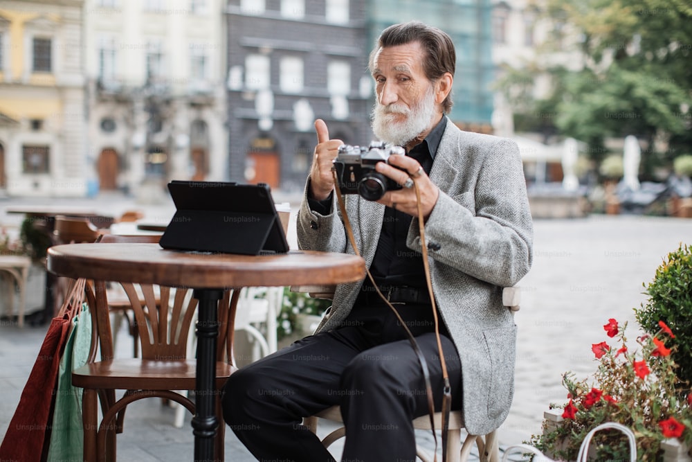 Confident senior man sitting at cafe terrace and using digital tablet with retro camera. Bearded pensioner reviewing pictures and showing thumb up.