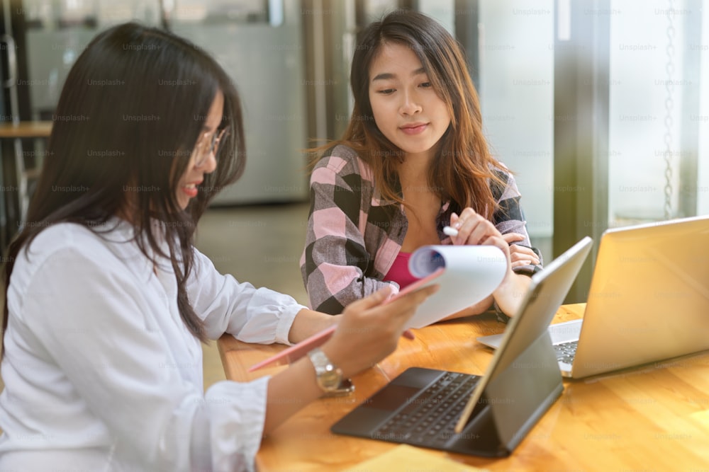 Side view of two young female university students consulting on their project with paperwork and two laptops