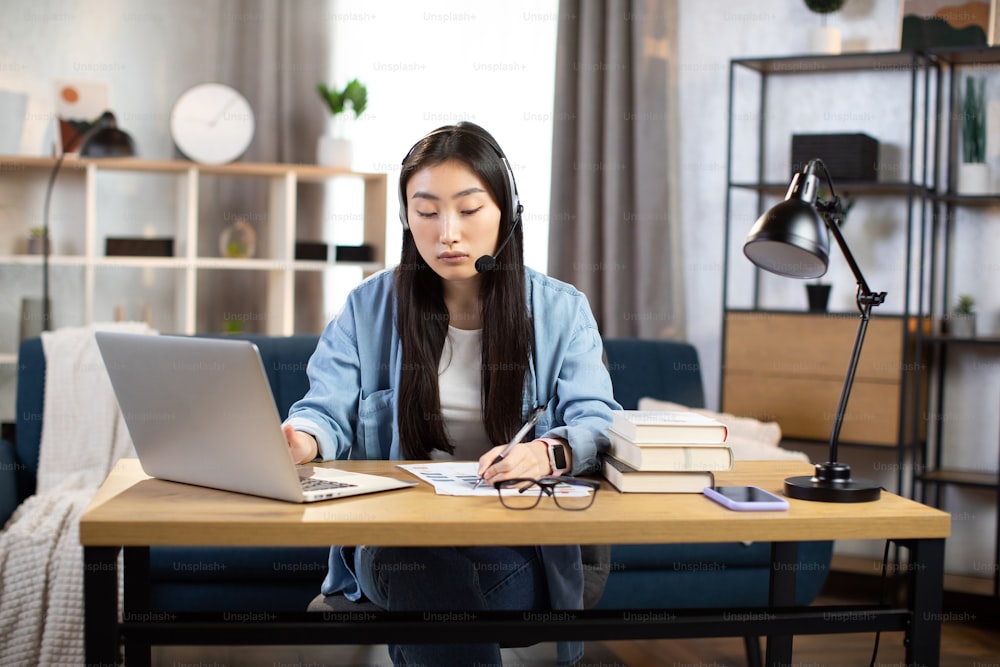 Mulher asiática nova usando fone de ouvido e laptop para conversa de vídeo no escritório. Freelancer feminina sentada à mesa e tomando notas durante o bate-papo por vídeo com o cliente em casa