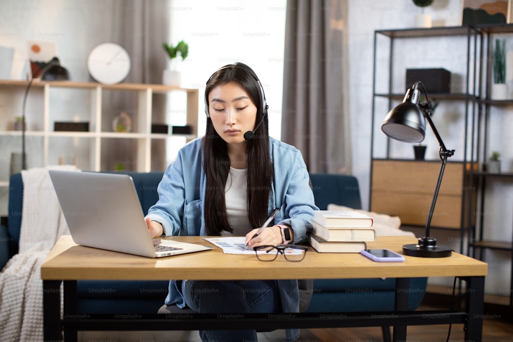 Young asian woman using headset and laptop for video conversation at office. Female freelancer sitting at table and taking notes during video chat with client at home