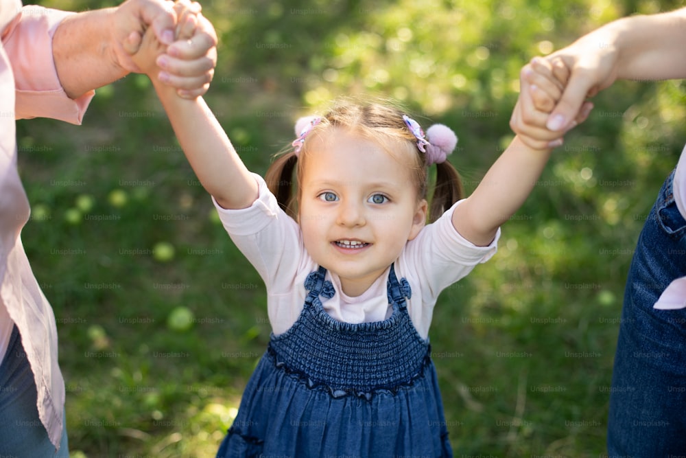 Top close up view of laughing adorable 3 years old child girl in jeans dress, having fun and enjoying her walk outdoors in the park, holding hands of her mother and grandmothe. Family walk in garden.